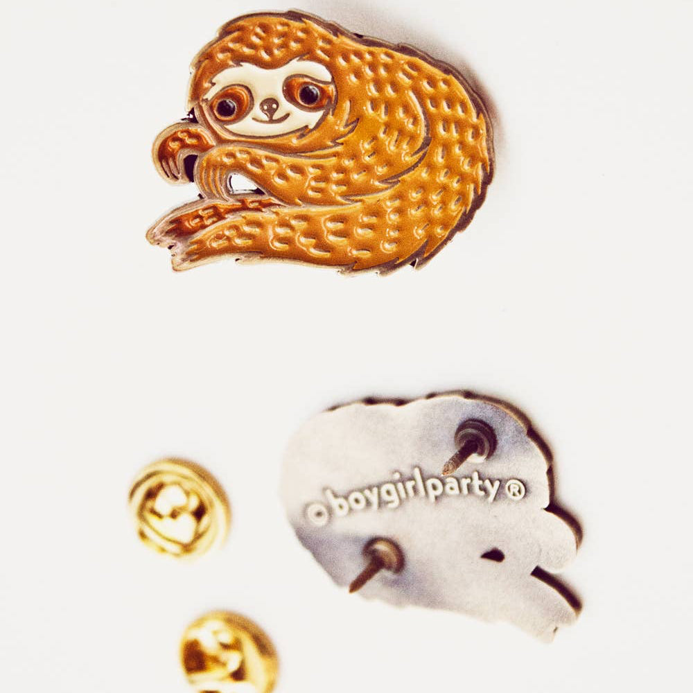 An enamel sloth pin by boygirlparty® is displayed on a white background with its backing and two gold post backings shown below. The sloth pin features a textured golden-brown body with a cream-colored face and eyes.