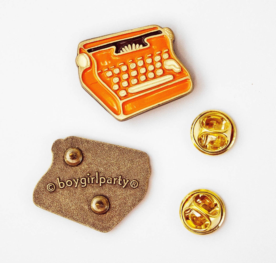 An enamel pin shaped like a typewriter in orange and gold, shown alongside its brown backing card and two gold-tone pin backs on a white background.