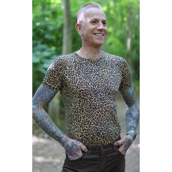 Image is of a person standing with their hands in their pockets, wearing a leopard print t shirt and black jeans.